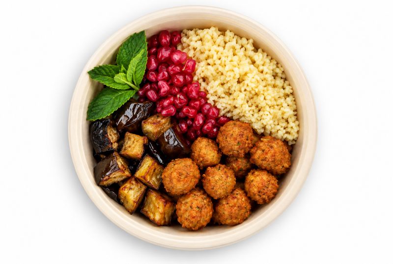 Vegane Bowl mit Couscous, Granatapfelkernen, gebratenen Auberginenwürfeln, goldbraunen Bällchen und Minze